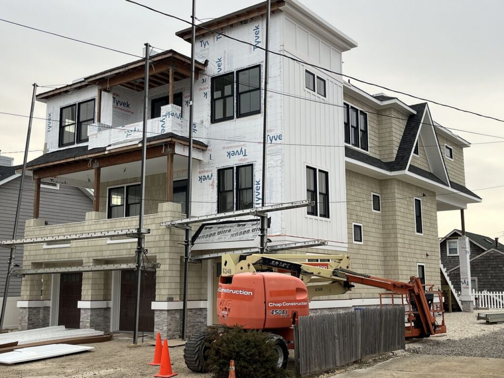 Chap Construction custom home build in progress in Seaside Park, New Jersey, showing a multi-story coastal residence with shake siding, stone veneer accents, and Tyvek house wrap still visible on the upper level, with a Chap Construction boom lift parked out front.