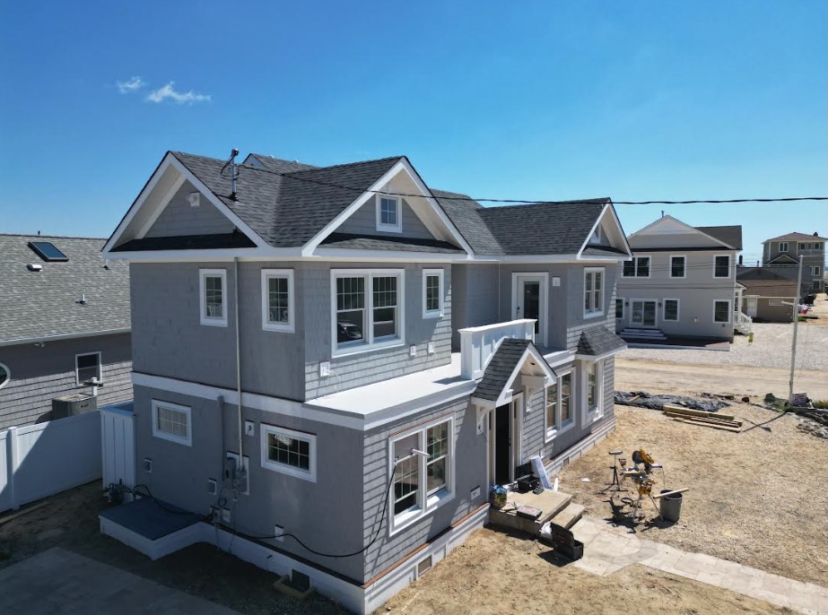 Exterior view of a bright, newly built room addition on a traditional two-story coastal New Jersey home featuring white vinyl siding, black-framed windows, a concrete foundation, landscaped flower beds, and a manicured lawn in a suburban neighborhood under natural daylight.