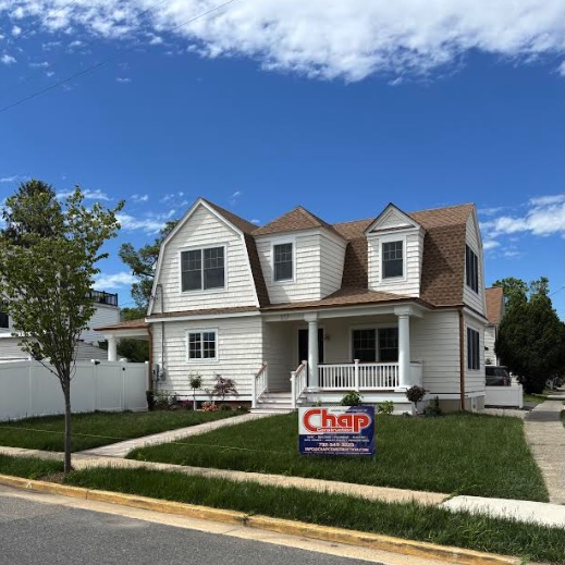 Two-story suburban home under construction with a second-story add-a-level addition in progress, featuring exposed wood framing, Tyvek house wrap, roofing underlayment, ladders, building materials, and construction workers on-site under a bright blue daytime sky.
